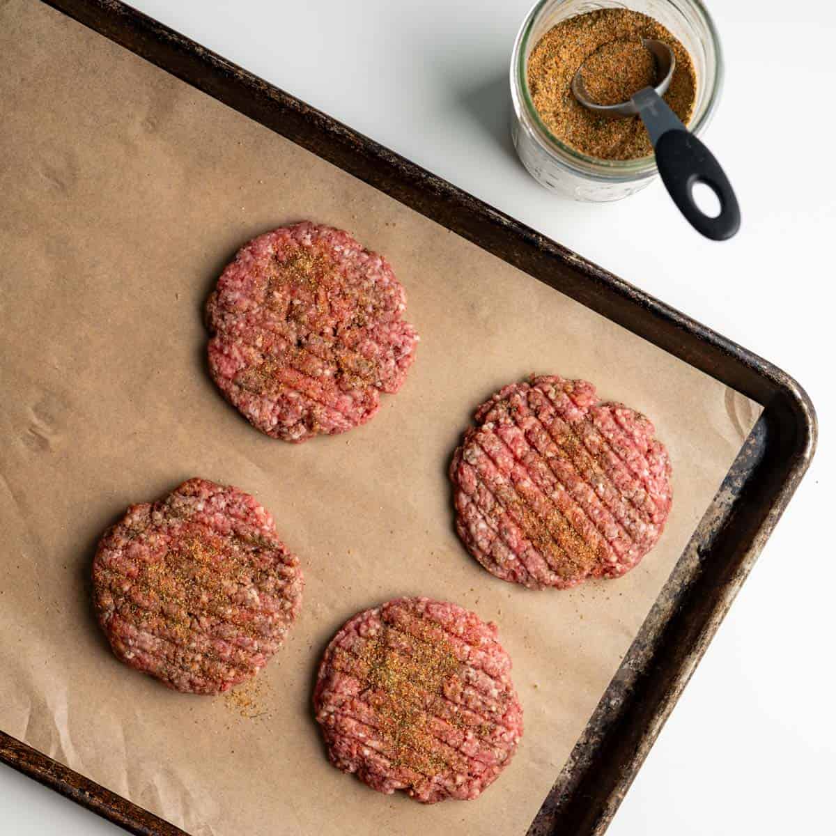 four uncooked burgers sprinkled with bloody mary seasoning on a baking sheet.