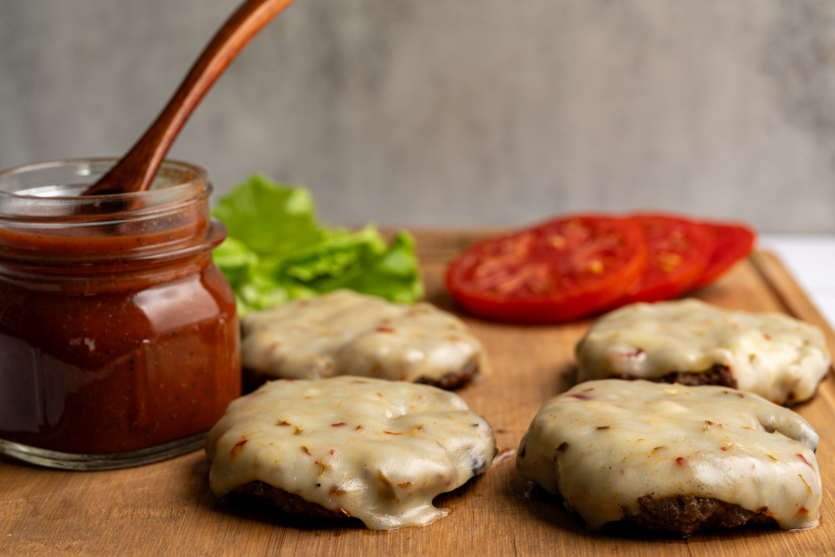four burgers covered with pepperjack cheese, lettuce, sliced tomatoes and bloody mary ketchup on a wooden board.