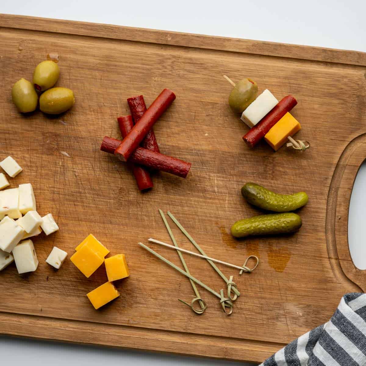 wooden cutting board with cubes of cheese, green olives, beef sticks and pickles.
