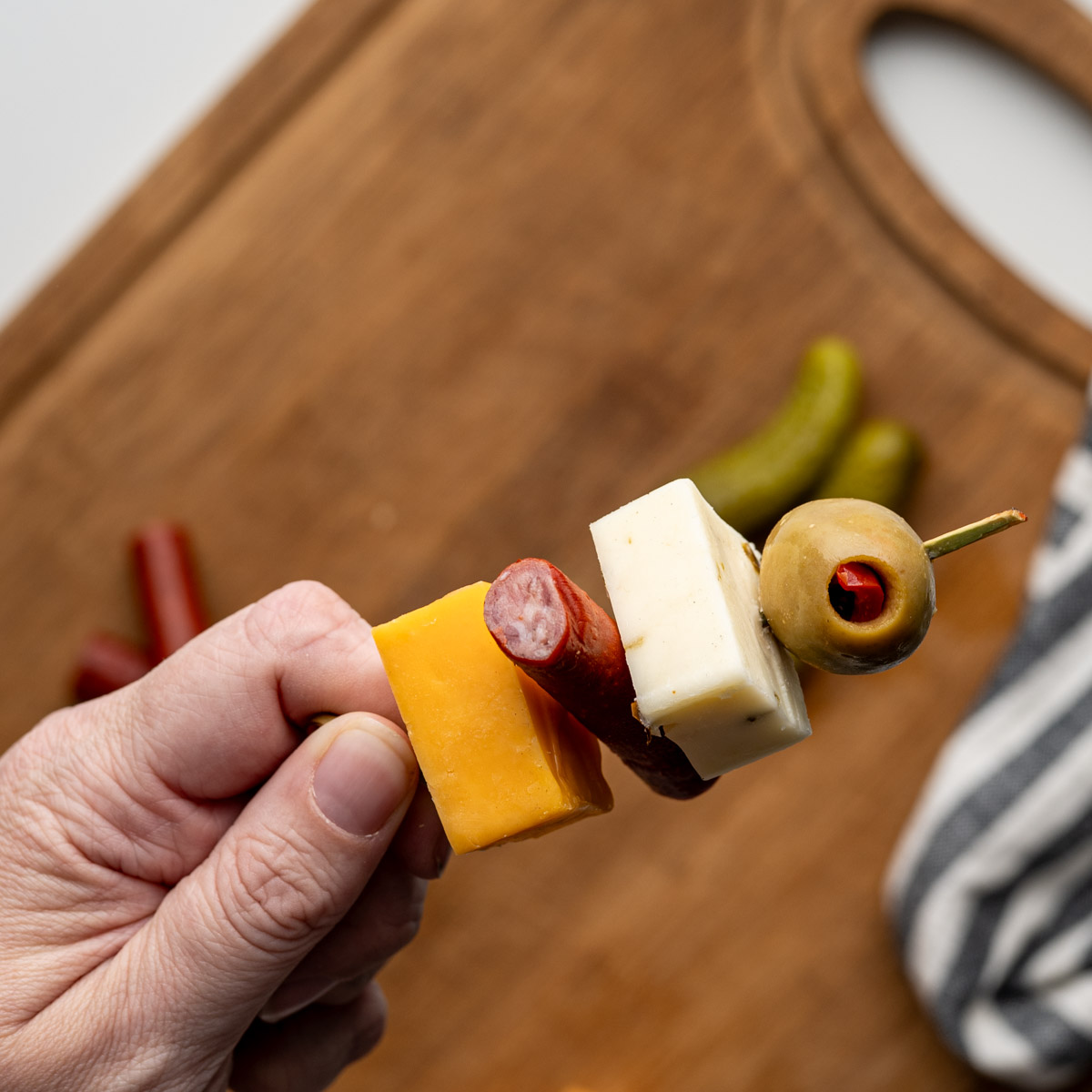 hand holding a toothpick with a green olive, cubed cheese and beef stick.