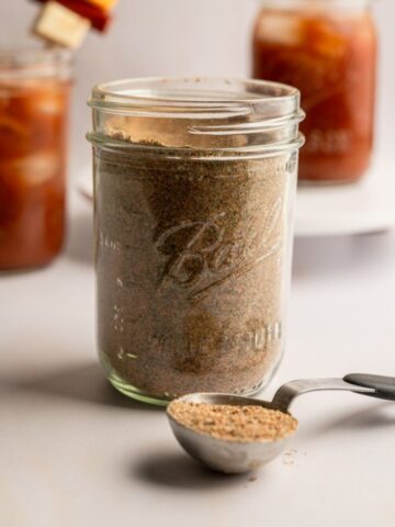mason jar filled with bloody mary seasoning with two bloody mary drinks in the background.
