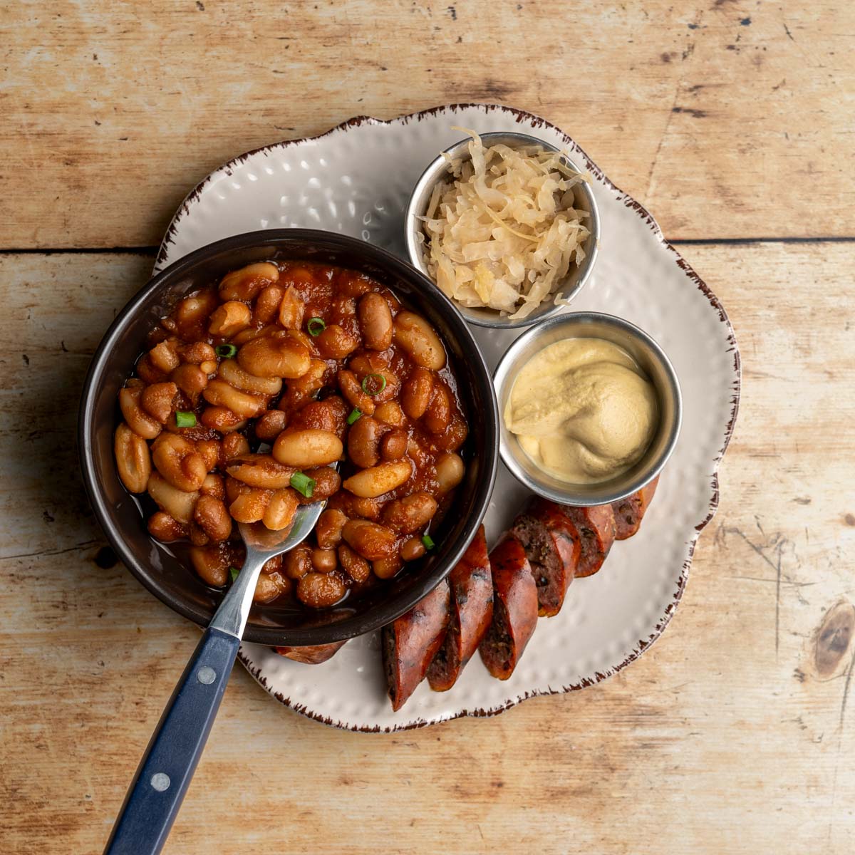 small bowl of baked beans on a plate with a sliced bratwurst, sauerkarut, and dijon mustard.