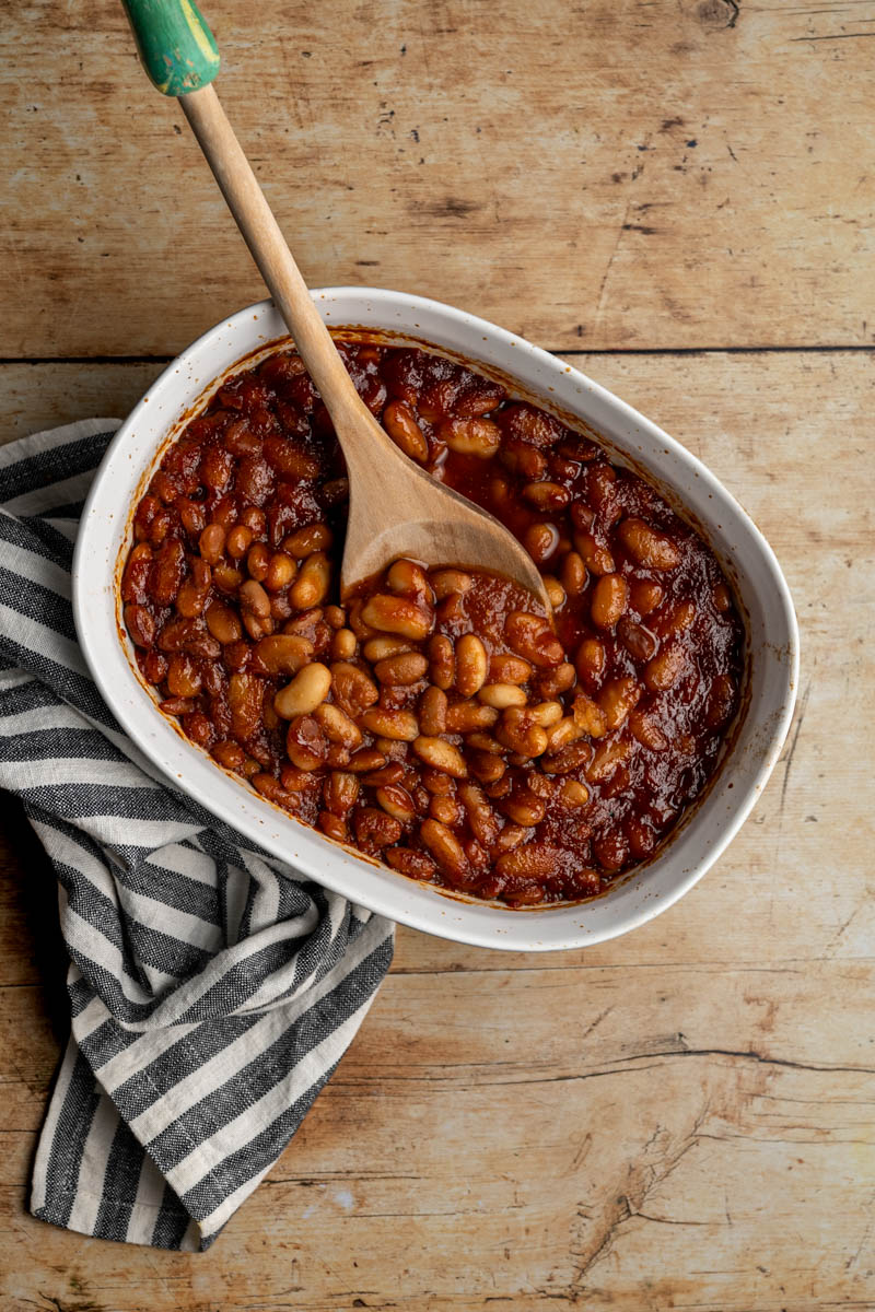 white baking dish filled with baked beans and a wooden spoon.