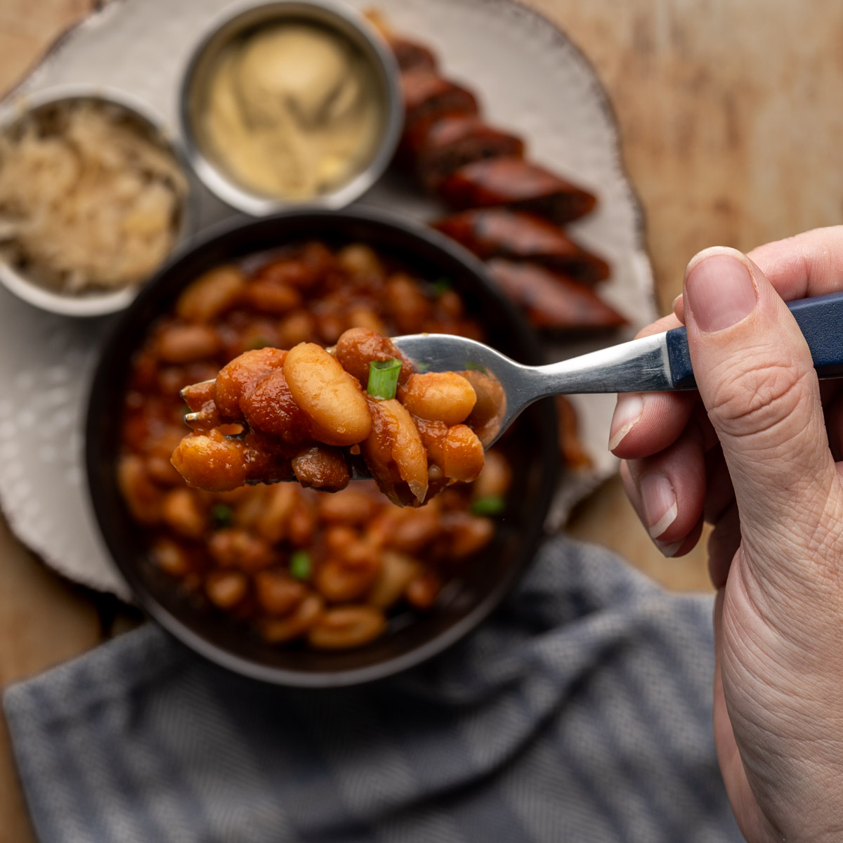 forkful of baked beans above a plate with a sliced bratwurst, sauerkraut and dijon mustard.
