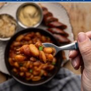 a forkful of baked beans about a plate with a sliced brat, sauerkraut, and dijon mustard.