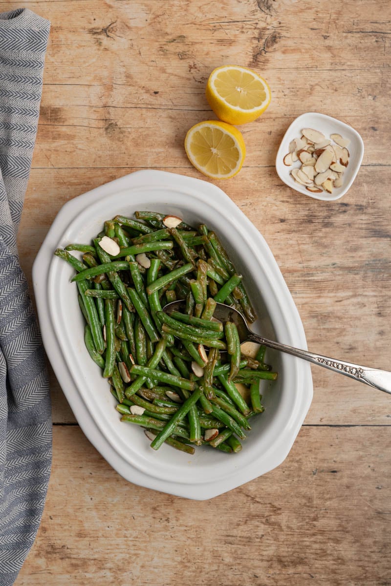 green beans in a white serving dish topped with silvered almonds, alongside a sliced lemon, sliced almonds.