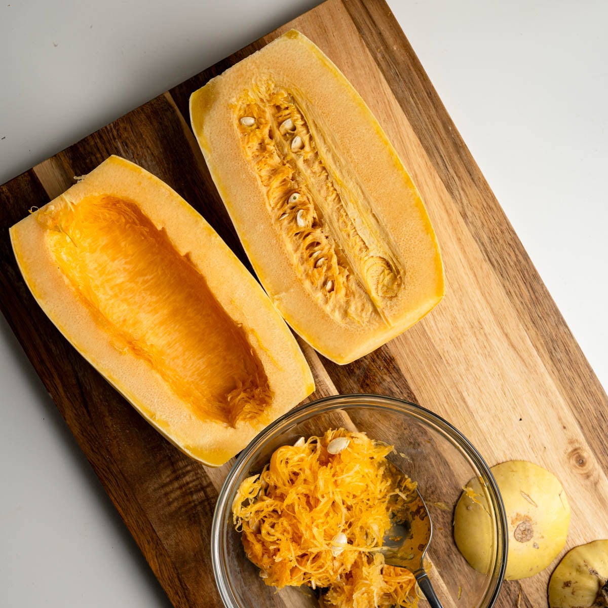 halved spaghetti squash, one deseeded with a glass bowl with seeds in it all on a wooden board.