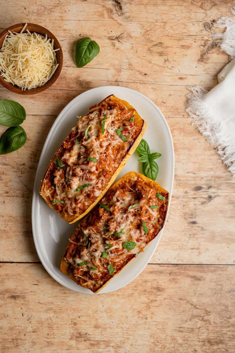 two stuffed spaghetti squash boats on a white platter topped with cheese, fresh basil on a wooden table.